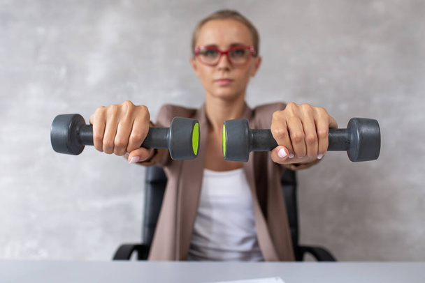 Blond woman in office clothes sits in chair and holding two dumbbells in hands above desk. Selective focus - Foto, Imagen
