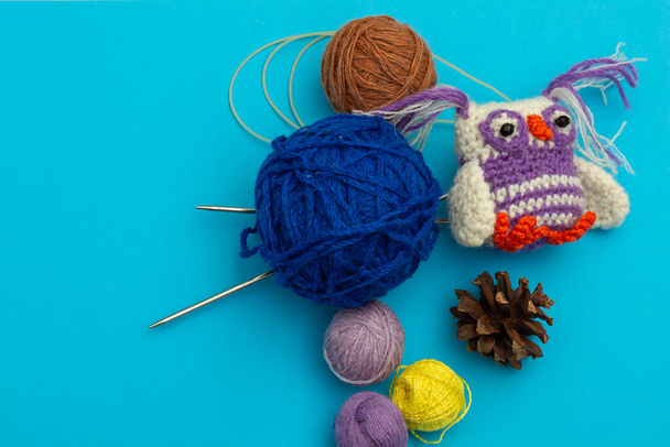 Petit jouet tricoté à la main coloré, hibou coloré à côté des écheveaux de fil pour la broderie, aiguilles à tricoter et cônes de pin secs sur un fond bleu. Le travail à l'aiguille et la créativité des enfants
 - Photo, image