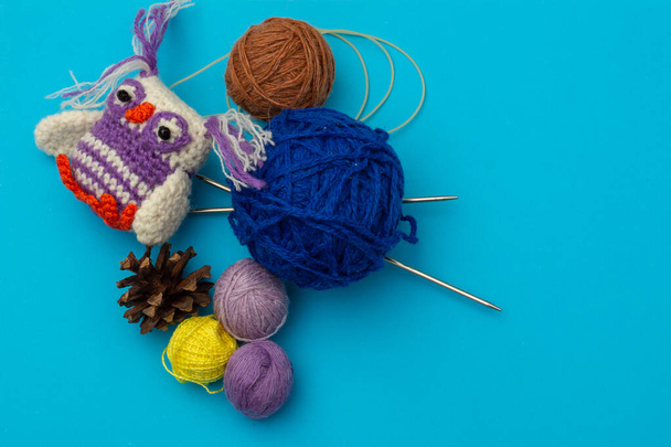 Neulottu käsintehty pieni värillinen lelu, värikäs pöllö lankojen vieressä käsityöhön, neulontaan ja kuiviin männynkäpyihin sinisellä pohjalla. Needlework ja lasten luovuuden käsite
 - Valokuva, kuva