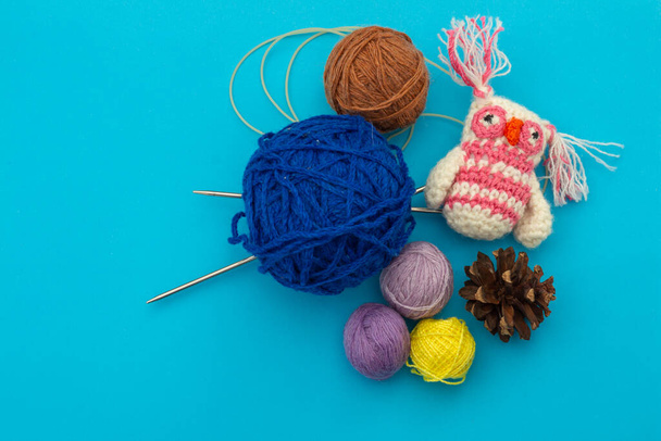 Petit jouet tricoté à la main coloré, hibou coloré à côté des écheveaux de fil pour la broderie, aiguilles à tricoter et cônes de pin secs sur un fond bleu. Le travail à l'aiguille et la créativité des enfants
 - Photo, image