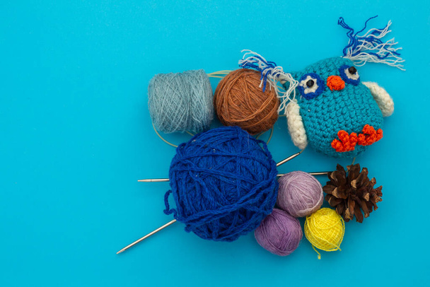 Knitted handmade small colored toy, colorful owlet next to skeins of thread for needlework, knitting needles and dry pine cones on a blue background.  Needlework and children's creativity concept - Photo, Image