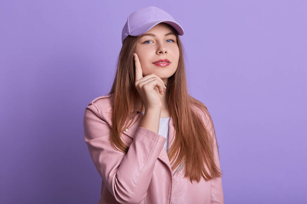 Closeup portrait of attractive lady with long blonde hair, dresses casual outfit, looking directly at camera, female keeping index finger on cheek, posing isolated over lilac studio background. - Photo, image
