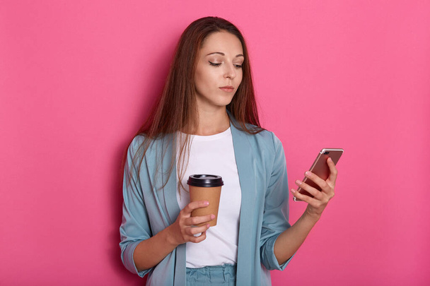 Horizontal shot of elegant concentrated female wearing suit, holding take away coffee and modern smart phone, woman looks confident and serious. Caucasian model poses isolate on pink studio background - Foto, Imagem