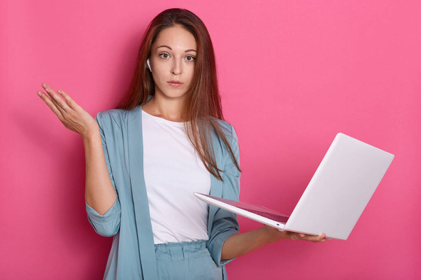 Closeup portrait of astonished and helpless woman posing with hand aside and holding white lap top in hand, lady looking directly at camera, posing isolated over pink studio background. Freelancer. - 写真・画像