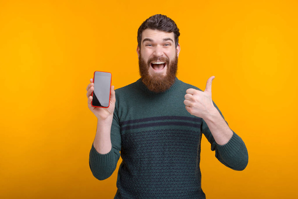 Exited bearded man is looking at the camera showing thumb up and his phone screen. - Fotó, kép