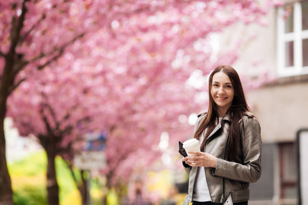 Girl in a great mood posing for a cup of coffee on Sakura background. Brunette woman near sakura - Foto, afbeelding