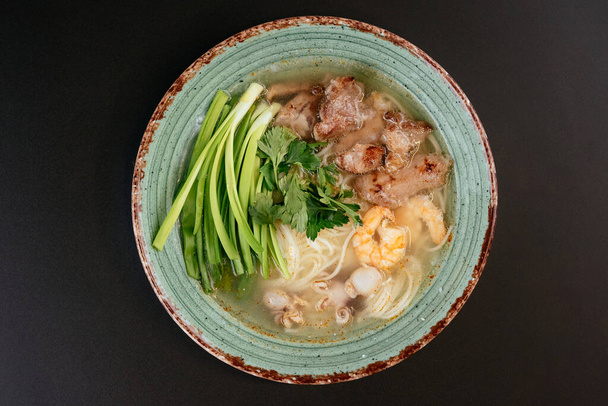 noodle soup with fried meat and shrimps with greens on green plate on black background  - Foto, Imagen