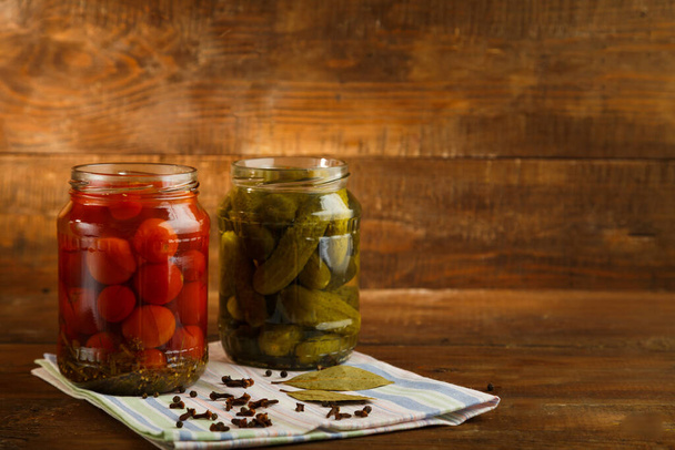 Jars of pickled homemade pickles and cherry tomatoes and sprinkled spices on a linen towel on a wooden table - Photo, Image