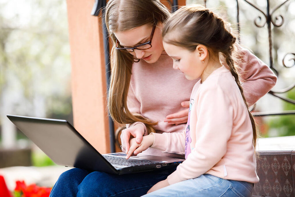 Молодая мать учит свою маленькую дочь печатать на ноутбуке, они сидят на заднем дворе дома
. - Фото, изображение