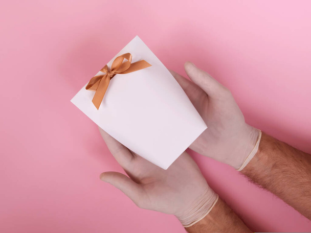 Αρσενικά χέρια σε ιατρικό γάντι κρατώντας μια λευκή τσάντα δώρου σε ροζ φόντο. Χωρίς επαφή δίνοντας ένα δώρο κατά τη διάρκεια της περιόδου καραντίνας. - Φωτογραφία, εικόνα
