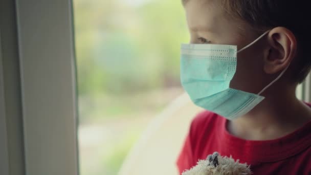 Sad illness child on home quarantine. Boy and his teddy bear both in protective medical masks sits on windowsill and looks out window. - Video