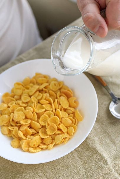 L'uomo ritagliato versa il latte in cornflakes in un piatto bianco su una tovaglia ruvida. Vista dall'alto. Concentrazione selettiva. Concetto, semplice veloce colazione americana sana
 - Foto, immagini