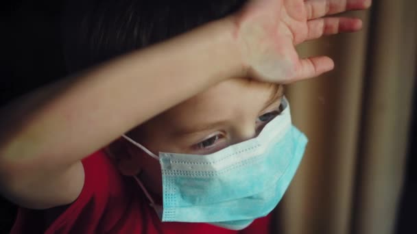 Ένα νεαρό αγόρι με ιατρική μάσκα κοιτάει έξω από το παράθυρο. απομόνωση σε καραντίνα, coronavirus, covid 19. - Πλάνα, βίντεο