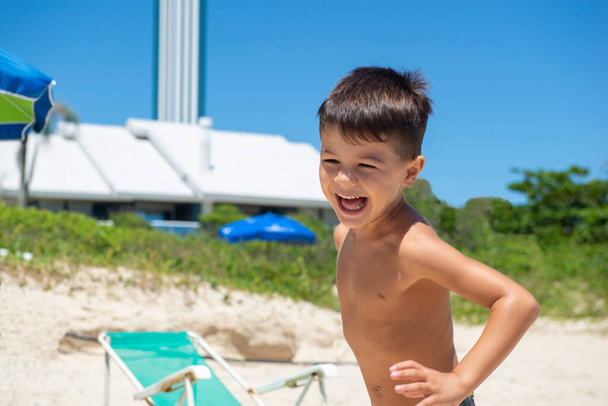 Kaukasischer Junge läuft und lächelt am Strand mit viel Grün im Hintergrund. Junge hat Spaß und spielt am Strand. Lächeln Sie entspannt und fröhlich. - Foto, Bild