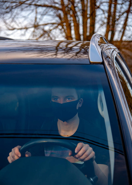 Ein junger Mann mit medizinischer Maske sitzt am Steuer eines Autos. - Foto, Bild