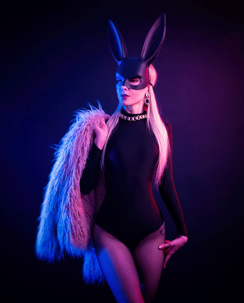 sexy girl in a bodysuit and fur coat with a rabbit mask in the neon light - Photo, Image