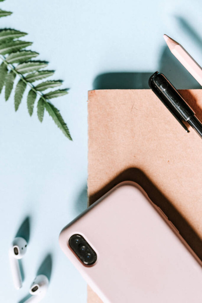A vertical flat lay picture of a smartphone, wireless earphones, notepad and pen. Pink case matches with blue background and green leaves. This modern and basic workspace is for nomads and millennial - Photo, Image
