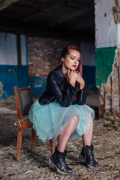 Young girl with pink hair dressed in leather jacket and a blue skirt sitting on the old chair inside of collapsed building surrounded by ruins. - Foto, afbeelding