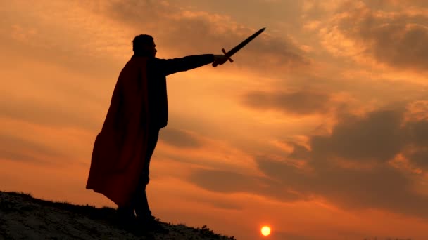 Özgür erkek şövalye. Elinde kılıçla ve kırmızı pelerinli Süpermen gün batımında bir dağın tepesinde duruyor. Özgür adam süper kahramanı oynuyor. Roma lejyonunun parlak güneş ışınlarıyla gökyüzüne karşı oyunu - Video, Çekim