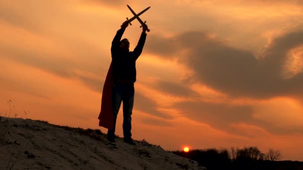 Ελεύθερος ιππότης διέσχισε τα σπαθιά του στον ουρανό πάνω από το κεφάλι του. Σούπερμαν με ένα σπαθί στο χέρι του και σε ένα κόκκινο μανδύα στέκεται στο βουνό στο φως του ηλιοβασιλέματος. Ο ελεύθερος άνθρωπος παίζει τον υπερήρωα. παιχνίδι της ρωμαϊκής λεγεώνας. - Πλάνα, βίντεο