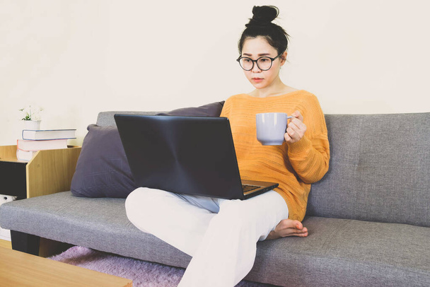 Молода красива азіатська жінка носила помаранчевий трикотаж, сиділа на сірому дивані, працювала онлайн-життям, відпочивала вдома з чашкою кави, домашнім робочим ноутбуком, працювала з дому
 - Фото, зображення