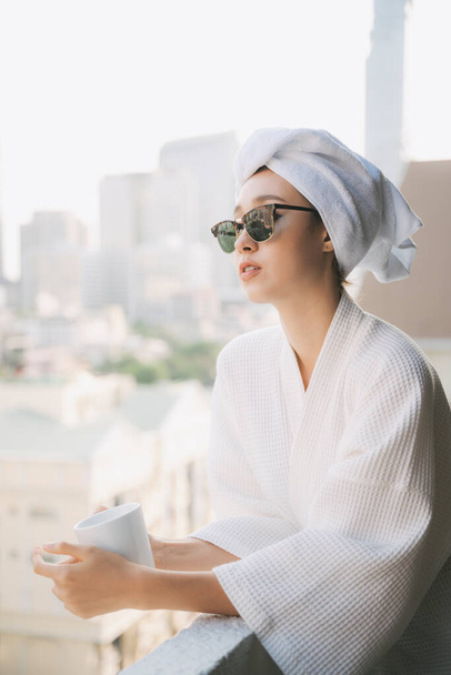 Mujer joven en albornoz con gafas de sol sosteniendo taza blanca de café en el balcón
. - Foto, imagen