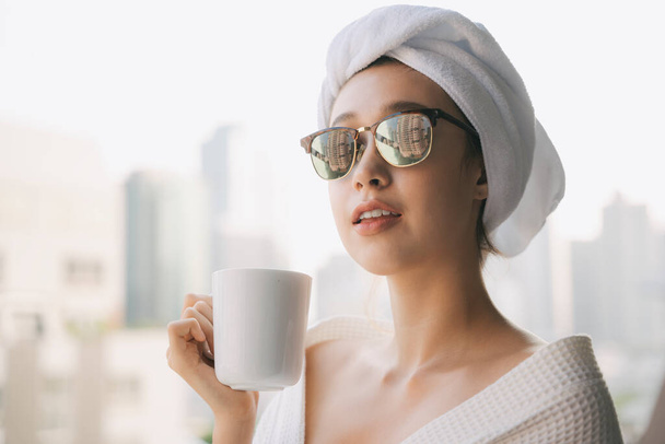 Close up portret - młoda kobieta w białym szlafroku i okularach przeciwsłonecznych gospodarstwa picia z filiżanki kawy na tle miasta. - Zdjęcie, obraz