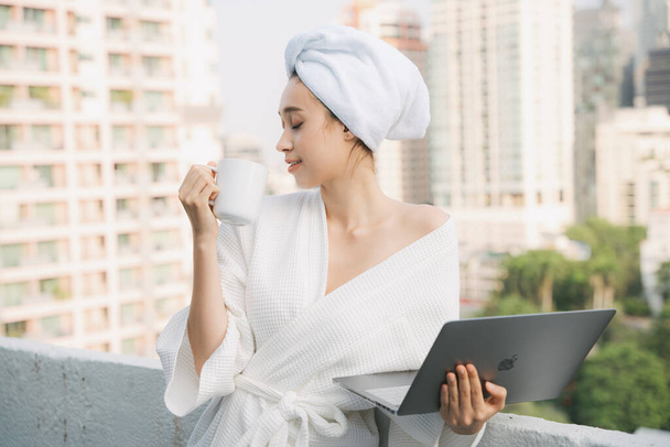 Young woman in white bathrobe enjoy hot coffee working at home with laptop in the morning. Work from home. - Photo, Image
