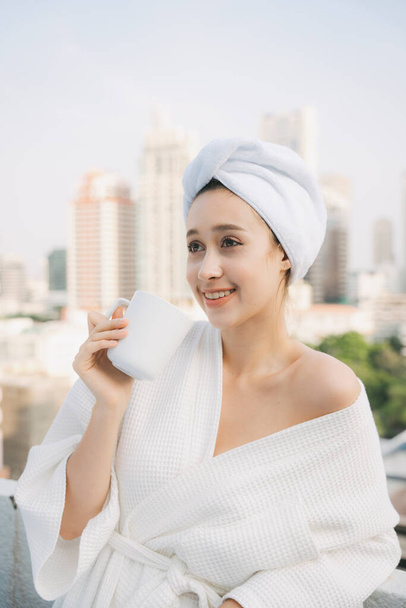 Junge Frau im weißen Bademantel genießt heißen Kaffee auf dem Balkon über der Stadt. - Foto, Bild