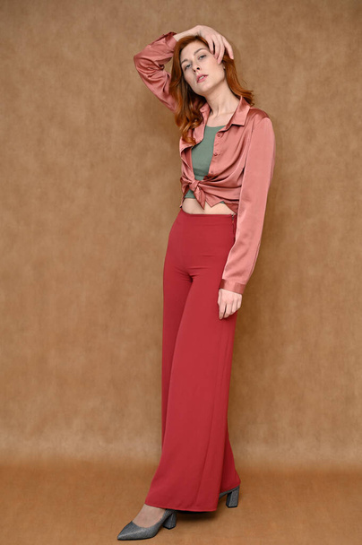 Vertical portrait of a red-haired woman in red trousers and a pink blouse is standing right in front of the camera. Photo taken on a beige background in the studio. - Photo, image