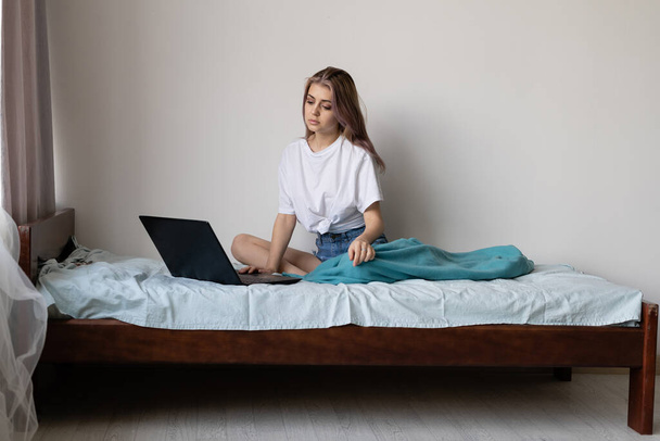 early morning a young beautiful woman is sitting on the bed. Women's clothing, white T-shirt. A laptop near female legs. Watch your favorite series. Chat with friends on social networks - Foto, Bild