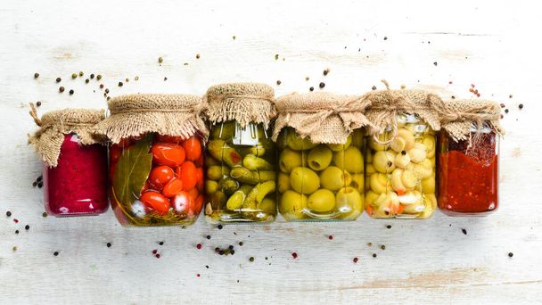 Canned food in glass jars on white wooden background. Pickled vegetables and mushrooms. - Фото, изображение