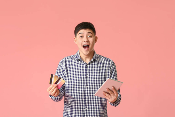 Surprised Asian man with credit cards and tablet computer on color background - Foto, Bild