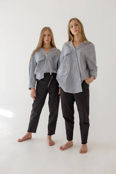 two young twin sisters in black jeans and striped oversize shirts with long blond hair posing on white background with bare feet and looking at camera. Stylish fashion casual clothes photoshoot - Foto, afbeelding