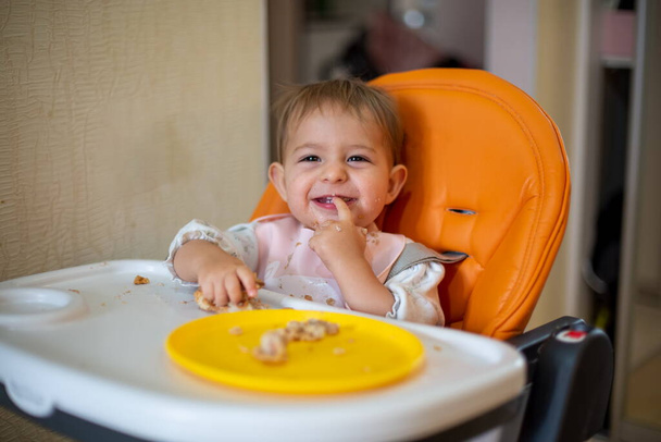 Cute baby w pomarańczowym foteliku dla dziecka patrzy na aparat śmiejąc się pokazując rękę na zębach.Zęby urosły. okruszki i pomarańczowy talerz z plackiem na stole. - Zdjęcie, obraz