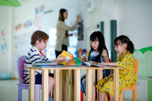group of international kids in preschool enjoy reading books with teacher watching in background - 写真・画像