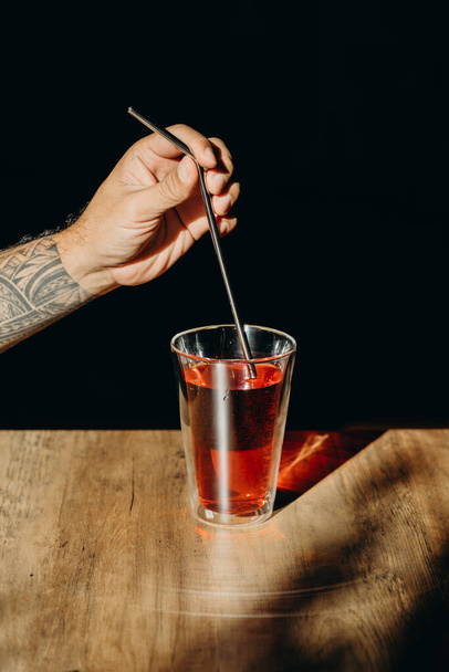 Una mano de hombre sosteniendo paja de metal o paja inoxidable por encima de un vaso de bebida dulce roja en la mesa de madera
. - Foto, Imagen