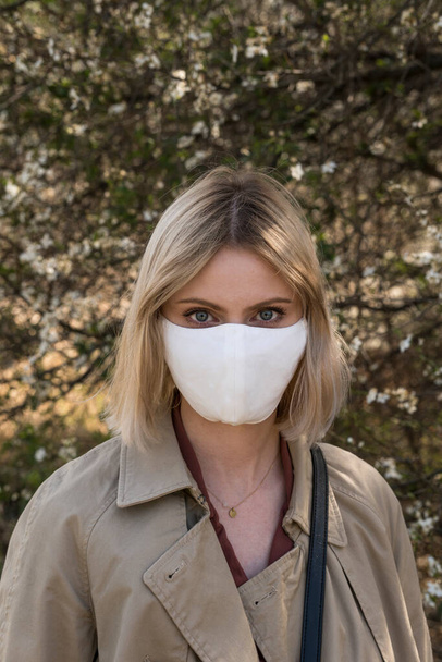 Jong blond meisje met een beschermend masker voor de verspreiding van het ziektevirus SARS-CoV-2. Epidemisch. Coronavirus. Covid-19. Bomen op de achtergrond. - Foto, afbeelding
