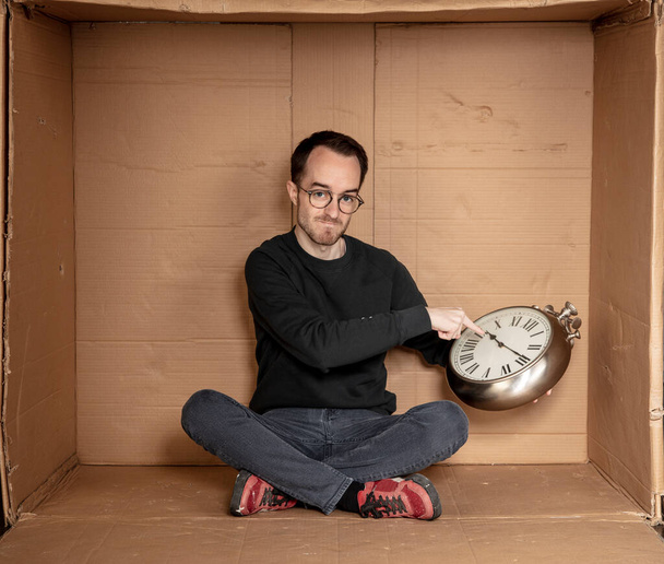 young unemployed student sits in a tight cardboard box, the idea of unemployment without experience, the clock in the hands of a symbol of the passage of time - Фото, изображение