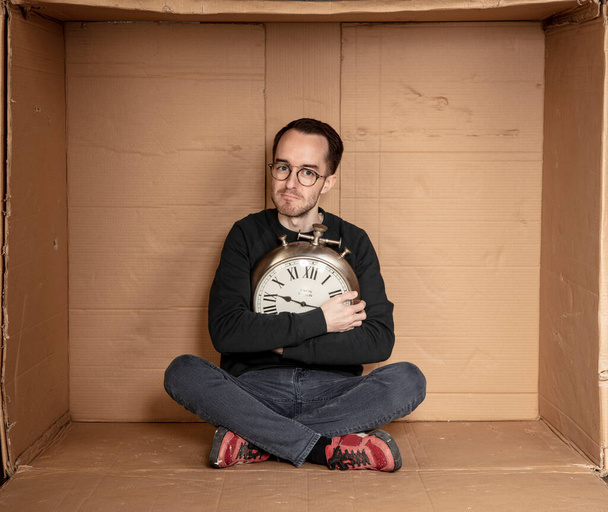 молодий безробітний студент сидить у щільній картонній коробці, ідея безробіття без досвіду, годинник в руках символу проходження часу
 - Фото, зображення