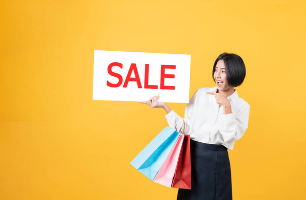 Joven mujer asiática sonriente sosteniendo bolsas de compras multicolores y manos mostrando letrero de venta sobre fondo naranja claro
. - Foto, imagen