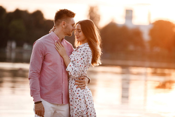 schöne verliebte Paar stehen und umarmen sich in der Nähe des Sees bei Sonnenuntergang. Schönes glückliches Paar, das Spaß im Freien hat. Glückliches Urlaubskonzept. - Foto, Bild