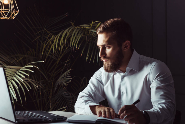 Handsome Thoughtful Bearded Businessman Sitting At His Workplace Looking At Laptop And Writing A Note. Business Theme - Foto, afbeelding