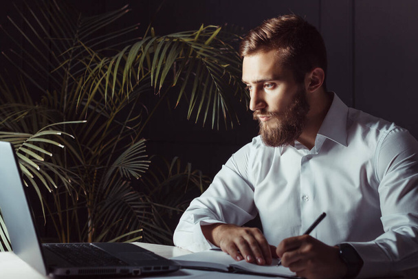Bonito pensativo barbudo empresário sentado em seu local de trabalho olhando para laptop e escrevendo uma nota. Tema de negócios
 - Foto, Imagem