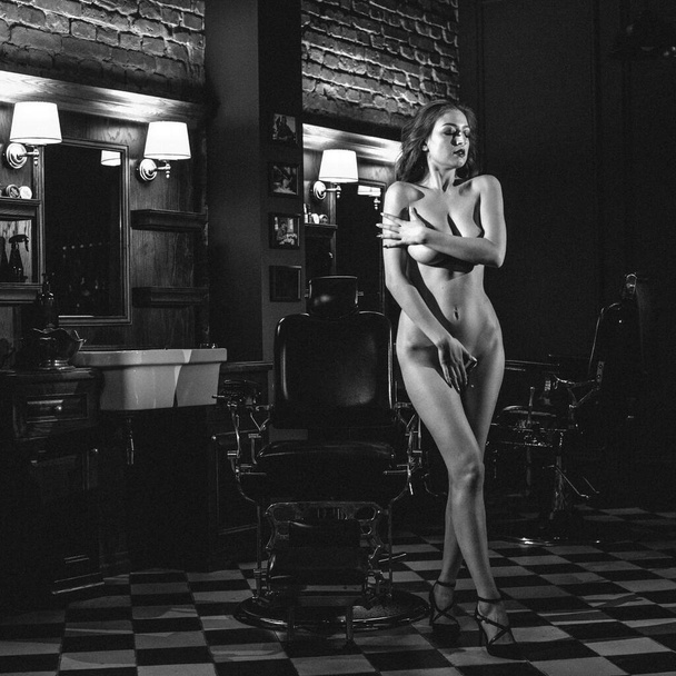 a young beautiful girl with good body shapes, naked, stands in a small room, in the center of which is a barber chair. evening light from lamps - Foto, imagen
