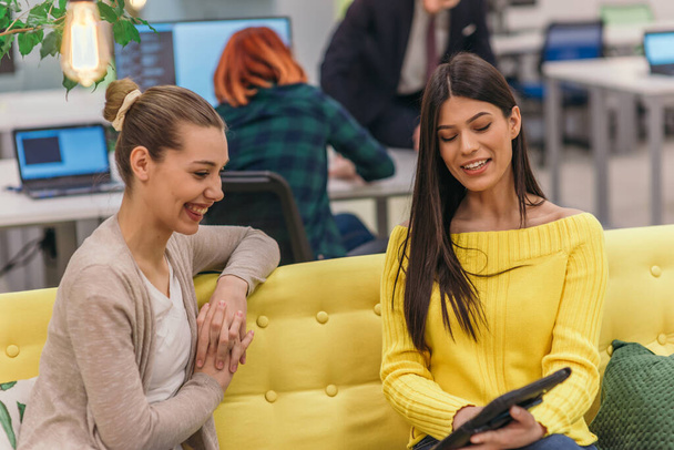 Multi-etnische collega 's blond blank en latina donker harig Knappe zakenvrouwen (werknemer) zitten op een gele bank terwijl ze werken op een laptop in een moderne startup bedrijf - Foto, afbeelding
