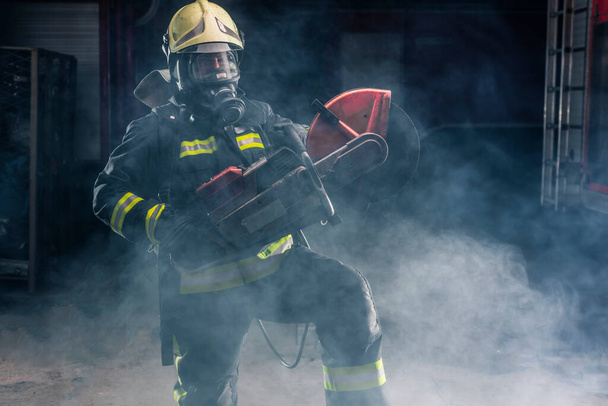 Portret van een brandweerman met beschermende uitrusting die poseert met een kettingzaag op zijn schouder. Donkere achtergrond met rook en blauw licht. - Foto, afbeelding