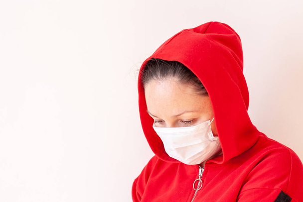 Een meisje met een medisch masker met een capuchon op haar hoofd liet haar hoofd zakken en kijkt naar beneden. Rode jas. Begrepen, ruimte. Witte achtergrond. Horizontaal. - Foto, afbeelding