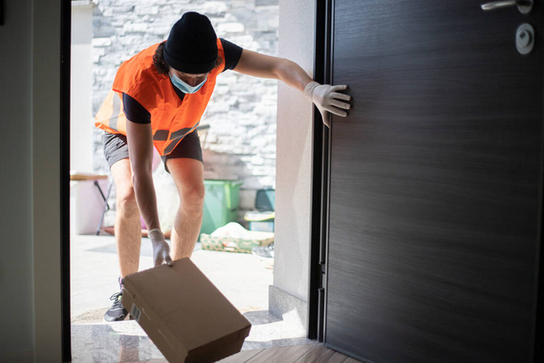 Young delivery man leaving a parcel box on the floor at front door, wearing gloves and face mask during covid-19 pandemic lockdown. Driver putting package on the floor just at open door, touching the door. - 写真・画像