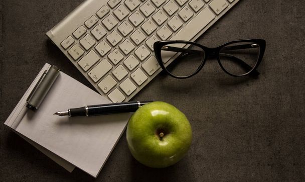 Objetos en la mesa de oficina gris oscuro. Gafas enmarcadas negras, teclado blanco, notebook y manzana verde
.  - Foto, imagen